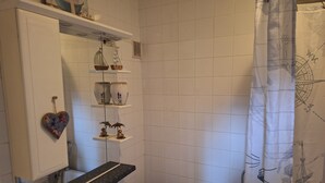 A bathroom with white tiles, a bathtub, a shower curtain with a compass design, and shelves with decorative items. - Private self catering apartment close to the stunning Pembrokeshire coast path. (Goodwick)