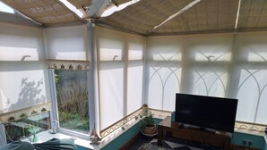 A room with a TV on a stand, a sofa, a window with a view of greenery, and a ceiling fan. - Private self catering apartment close to the stunning Pembrokeshire coast path. (Goodwick)