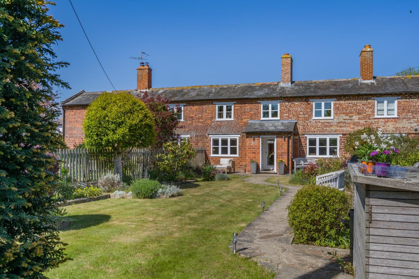 Une maison à deux étages, construite en briques, avec un jardin devant, une clôture en bois et un chemin menant à l’entrée.
