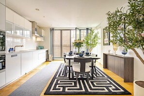 A modern kitchen with white cabinets, a dining area with a black and white rug, and a large window with a view of a brick building. - The Fitzrovia Grand Residence (London)