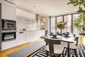 A modern kitchen with white cabinetry, a dining area with a table set for four, and a large window with a view of a brick building. - The Fitzrovia Grand Residence (London)