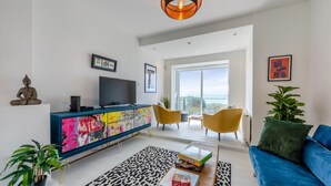 A modern living room with a blue sofa, a colorful cabinet, a television, and a large window offering a view. - 4 Bed in Saltdean (oc-a32366) (Saltdean)