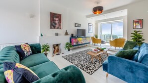 A modern living room with a sofa, armchairs, a coffee table, and a TV. There is a fireplace, a large window, and a balcony with a view. - 4 Bed in Saltdean (oc-a32366) (Saltdean)