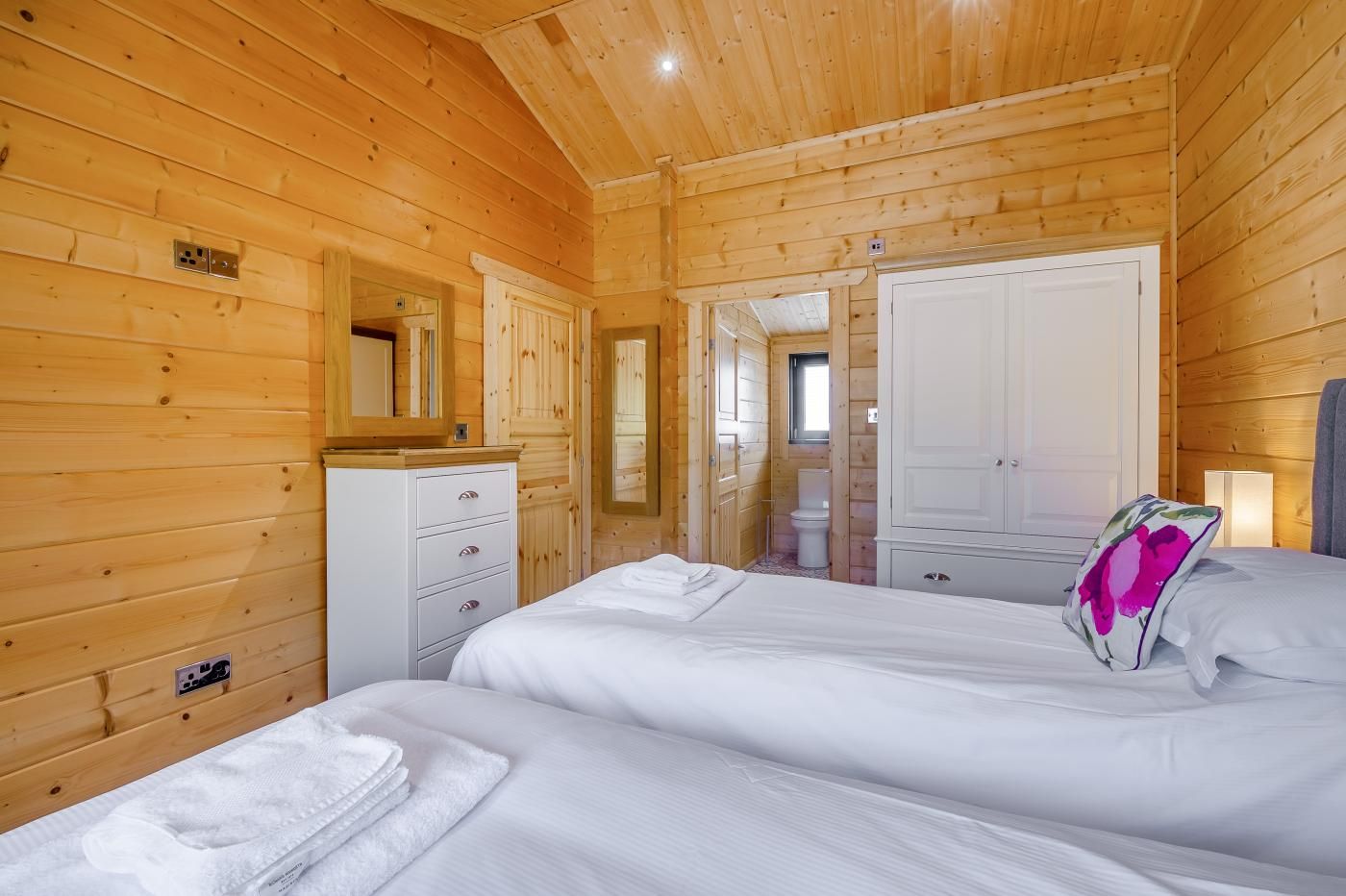 A wooden cabin bedroom with two beds, a dresser, a mirror, and a door leading to a bathroom.