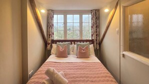 A bedroom with a large bed, a window with curtains, and a wooden ceiling beam. - 17th Century Cottage | Suffolk Market Town (Hadleigh)