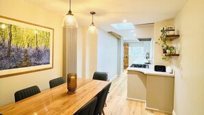 A modern dining area with a wooden table and chairs, pendant lighting, and a kitchen in the background. - 17th Century Cottage | Suffolk Market Town (Hadleigh)