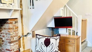 A staircase with white railings, a brick fireplace, a white cushioned chair with floral upholstery, and a wooden cabinet with a television on top. - 17th Century Cottage | Suffolk Market Town (Hadleigh)