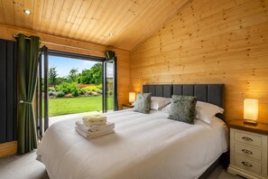 Ein Schlafzimmer mit Bett, Nachttischlampen, Blick auf einen Garten und einer Holzbalkendecke.