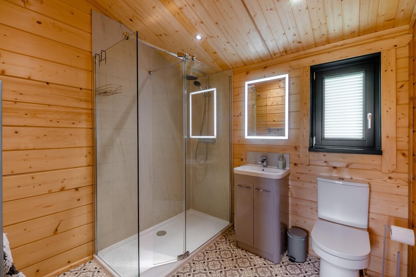 Ein Badezimmer einer Holzhütte mit einer Duschkabine aus Glas, einem Waschbecken, einer Toilette und einem Fenster mit Jalousien.