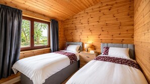 A wooden cabin room with two beds, a window with curtains, and a nightstand with a lamp. - 2 Bed in Llandinam (oc-t31974) (Llandinam)