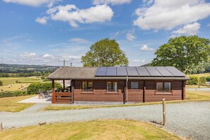 Eine Holzhütte mit geneigtem Dach und Solaranlage, umgeben von einer Schotterauffahrt und einer Rasenfläche mit einer Bank.