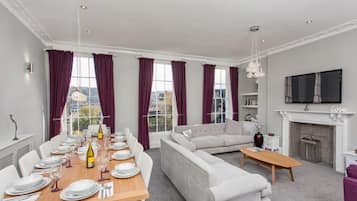 A modern living room with a dining area, white sofas, a fireplace, and a TV mounted on the wall.