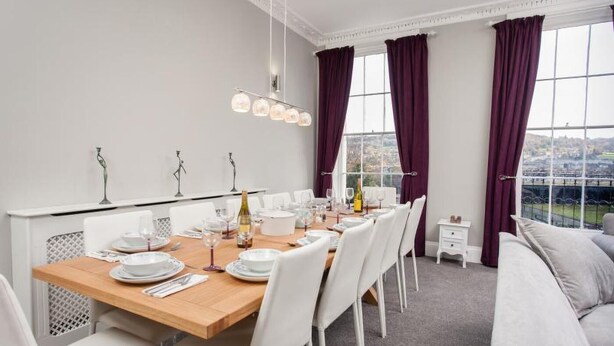 Comedor con una mesa de madera dispuesta para cuatro personas, sillas blancas y vistas a un paisaje urbano a través de ventanas amplias con cortinas moradas.