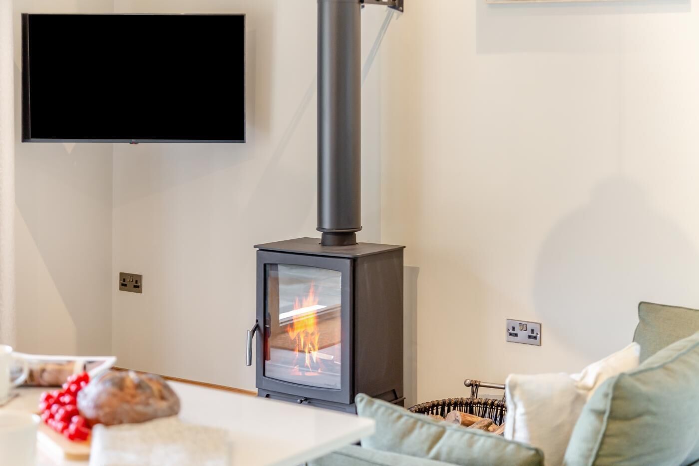 A modern living room with a wood-burning stove, a flat-screen TV mounted on the wall, and a sofa with cushions.