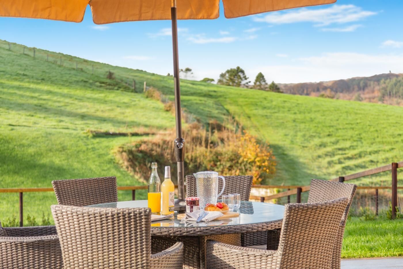 A table with wicker chairs and an umbrella, set against a backdrop of rolling green hills.