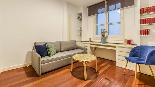 Ein Zimmer mit einer Couch, einem kleinen runden Tisch, einem Stuhl, einem Fenster und einem Bücherregal.