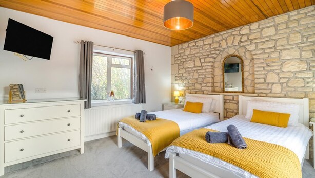 A room with two beds, a stone wall, a wooden ceiling, a flat-screen TV, a dresser, and a window with curtains. - 4 Bed in Bath (oc-bhr4) (Bath)