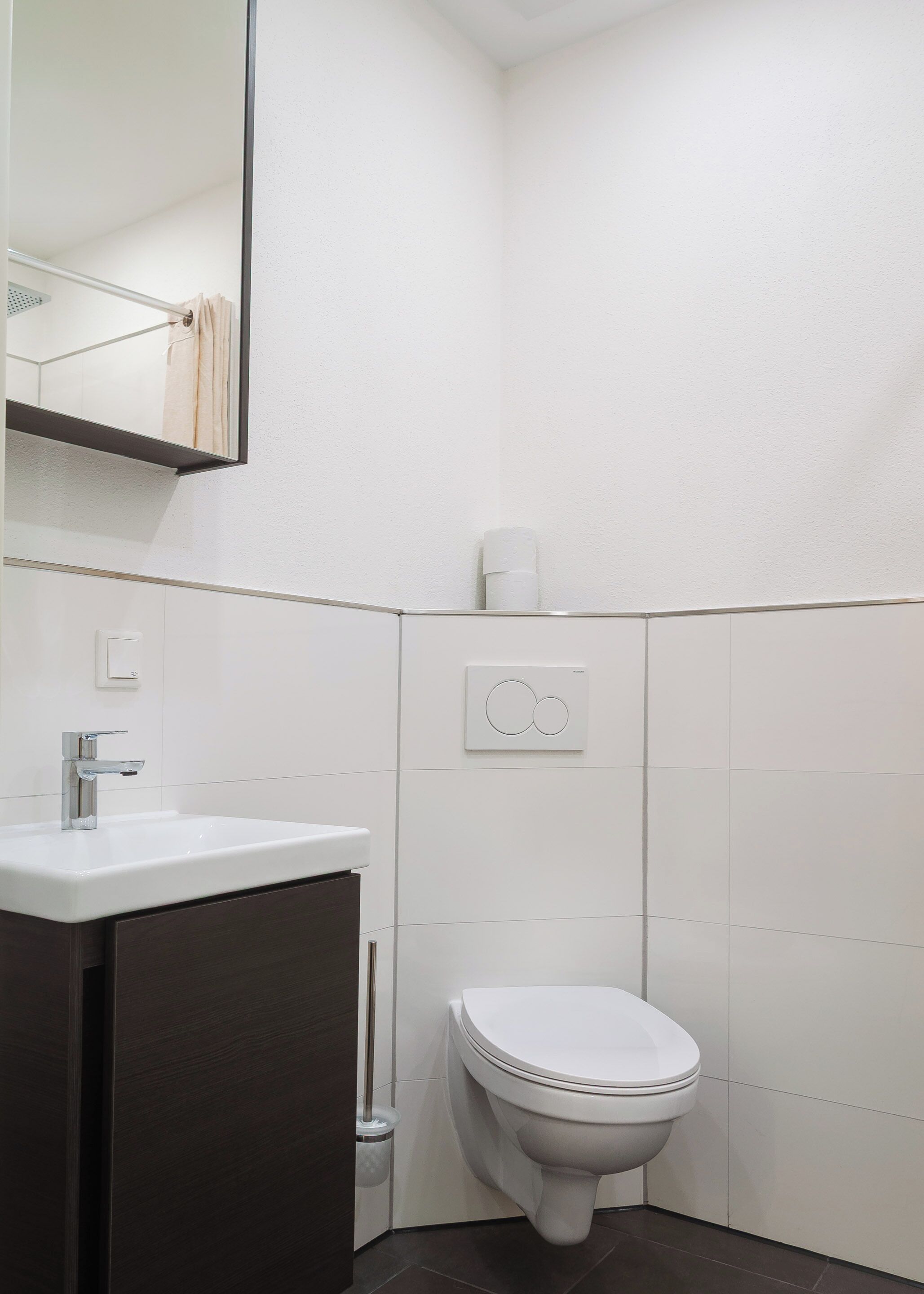 Ein modernes Badezimmer mit Waschbecken in Weiß, einem dunklen Schrank, einer Toilette und einem Spiegel.