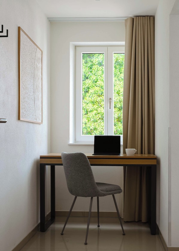 A room with a desk, a laptop, a chair, a window with curtains, and a framed picture on the wall.