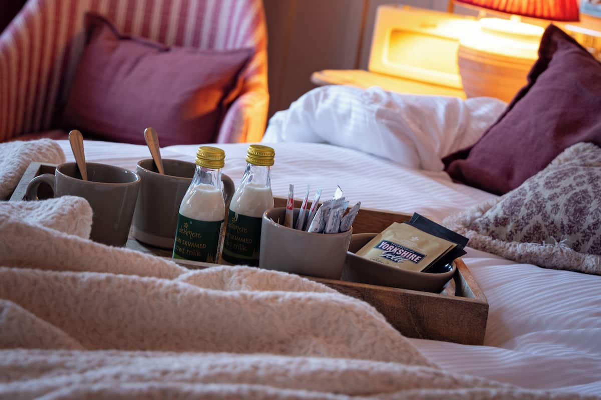 Habitación de hotel con cama, mesita de noche, lámpara y una bandeja con botellas de leche y una tarjeta.