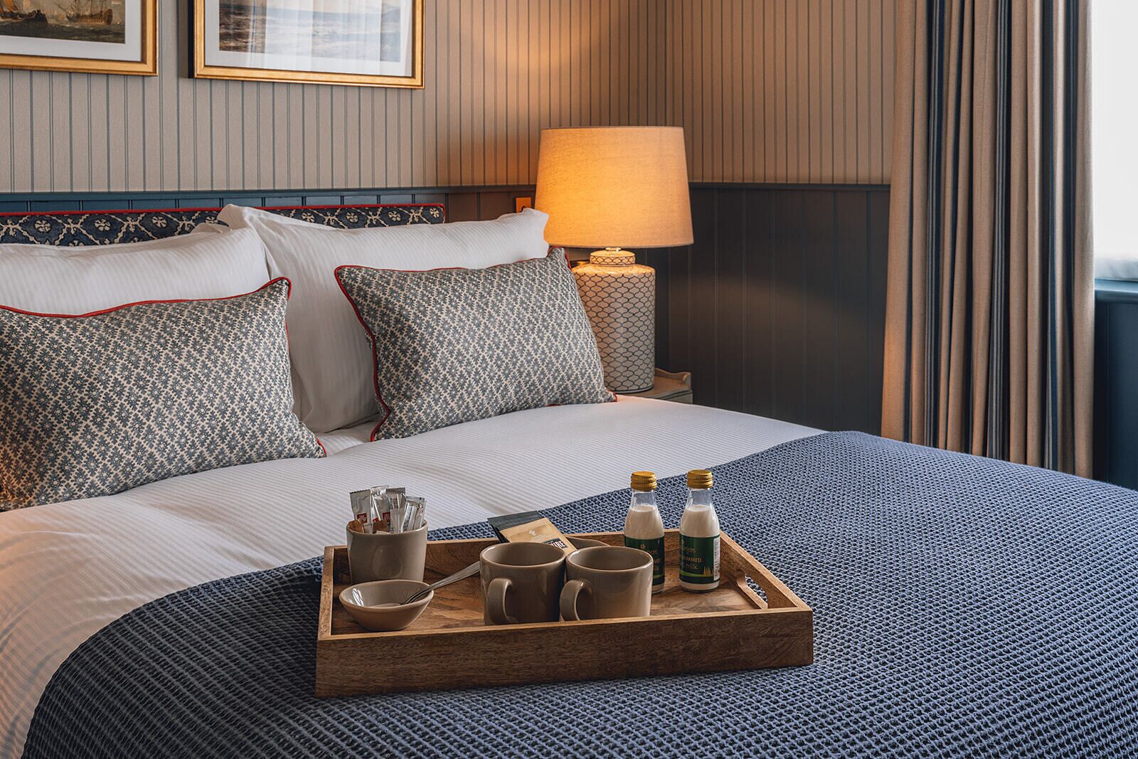 A neatly made bed with patterned pillows, a bedside table with a lamp, and a tray with breakfast items.