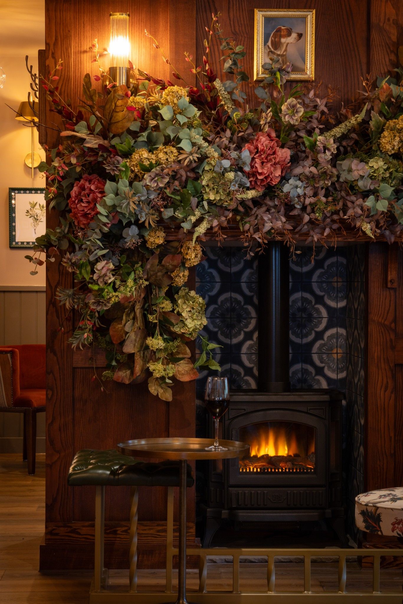 A fireplace with a decorative floral arrangement above it, a glass of red wine on a table, and a framed picture on the wall.