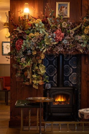 Una chimenea con un arreglo floral decorativo encima, una copa de vino tinto sobre una mesa y un cuadro en la pared.