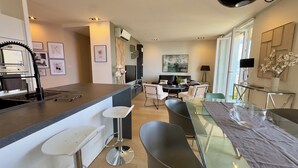 Una cocina moderna con una isla central, taburetes de bar, un comedor con mesa de vidrio y una sala de estar con sofá y mesa de centro.