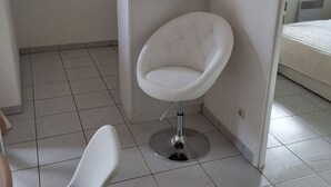 A small, modern kitchen with a white chair, a small table, and a bed in the adjacent room.