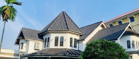 Una casa grande de varios pisos con una torre central destacada, rodeada de un césped cuidado y diversas plantas en macetas.