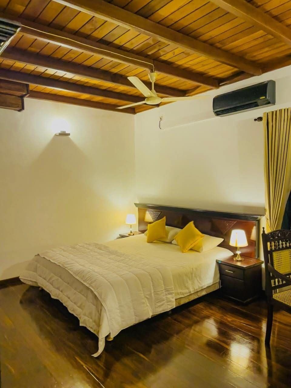 A hotel room with a wooden ceiling, a bed with white bedding, two bedside lamps, and an air conditioning unit.