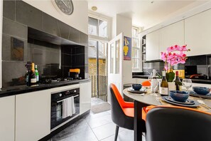 A modern kitchen with black and white cabinetry, a gas stove, and a dining area with orange chairs and a table set for two. - The Marylebone Maison - 4 Bed Apartment with AC (London)