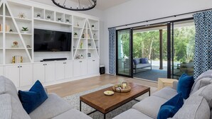 Ein modernes Wohnzimmer mit großem Fernseher, weißen Regalen und einer Glastür, die zu einem Außenbereich führt.