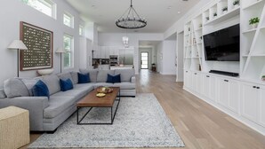 Ein geräumiges Wohnzimmer mit einem grauen Sofa, einem Couchtisch aus Holz und einem großen Fernseher auf einer weißen Schrankwand.