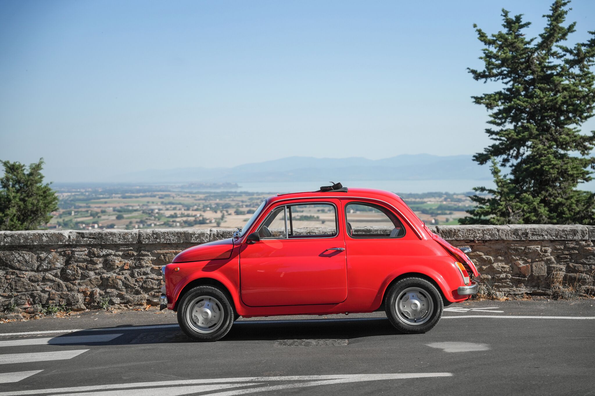 Ein rotes Auto parkt an einer Straße mit einer Steinmauer und einem schönen Ausblick auf ein Tal und Berge im Hintergrund.