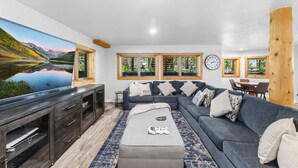 Un salon moderne doté d’un grand téléviseur à écran plat, d’un canapé d’angle gris, d’un meuble TV en bois et d’une grande fenêtre offrant une vue sur les montagnes.