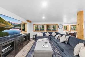 A modern living room with a large flat-screen TV, a grey sectional sofa, a wooden TV stand, and a large window with a mountain view. - Breckenridge Outdoor Oasis | Pool Table + Hot Tub (Breckenridge)