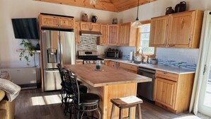 A modern kitchen with wooden cabinets, stainless steel appliances, a large island with stools, and a hanging light fixture. - Entire Home in Carthage, ME (Carthage)
