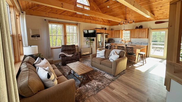 A spacious living area with a wooden ceiling, a sofa set, a coffee table, and a kitchen with wooden cabinets and a large window. - Entire Home in Carthage, ME (Carthage)