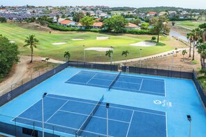 Ein Tennisplatz mit zwei Spielfeldern, umzäunt, mit einem Golfplatz im Hintergrund.