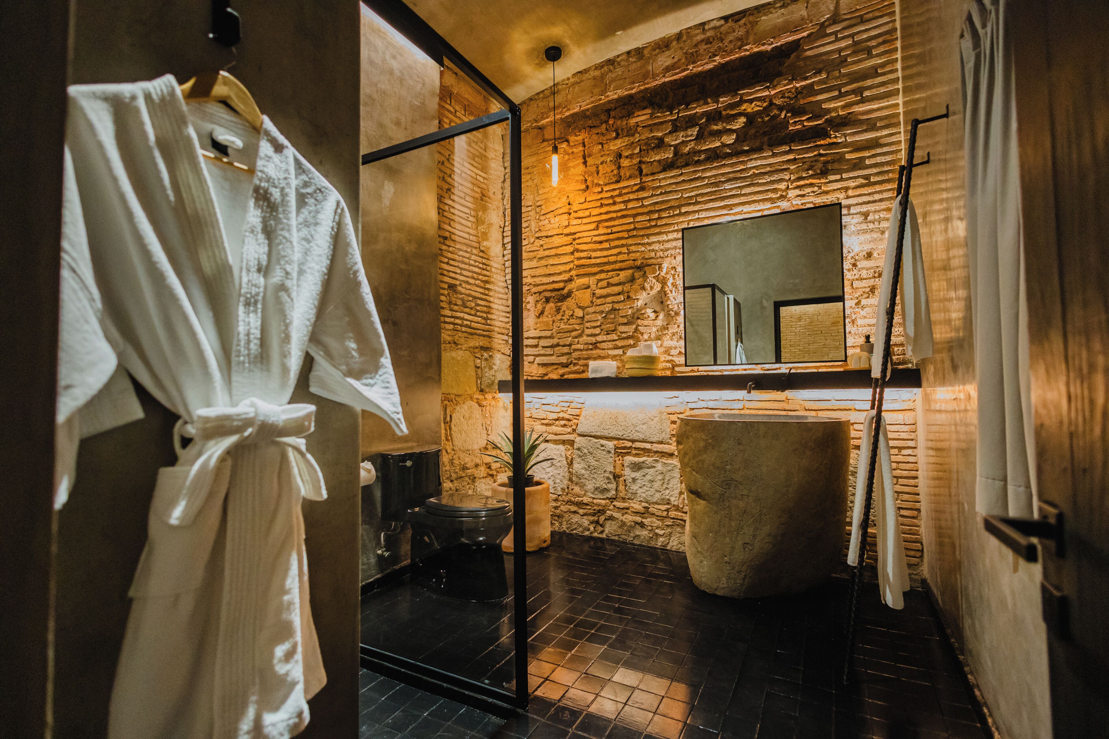 A bathroom with a stone wall, a large mirror, a bathtub, and a robe hanging on a rack.