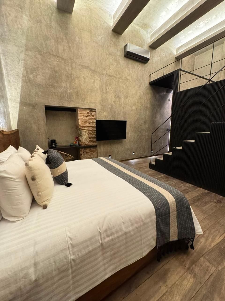 A bedroom with a large bed, a flat-screen TV mounted on the wall, a fireplace, and a staircase.