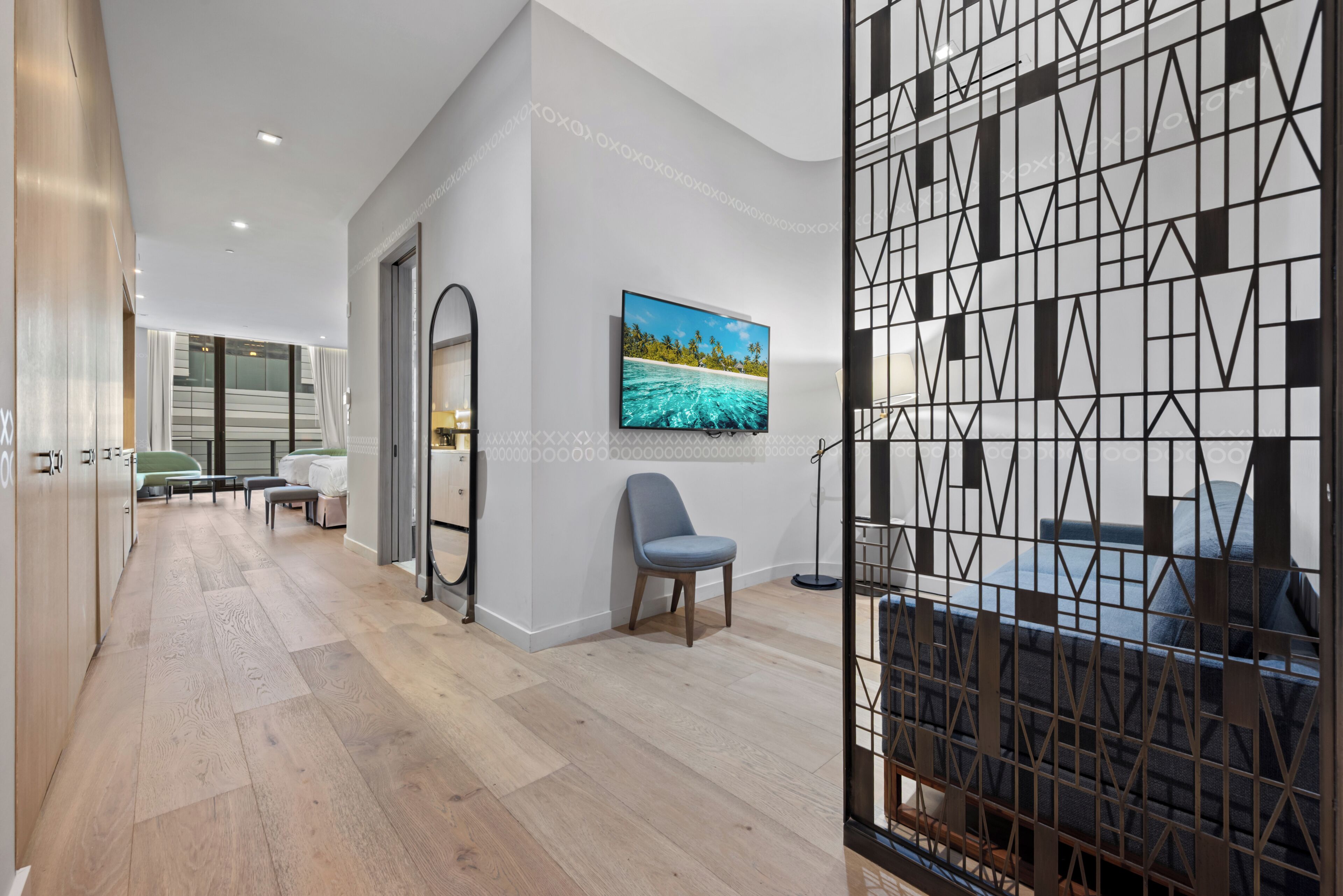 Een moderne hotelkamer met een houten vloer, een groot raam, een schilderij van een strand en een geometrisch wandontwerp.