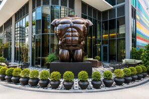 Ein modernes Gebäude mit einer großen Bronzeskulptur einer muskelbepackten Figur in der Mitte, umgeben von Topfpflanzen und Außenbestuhlung.