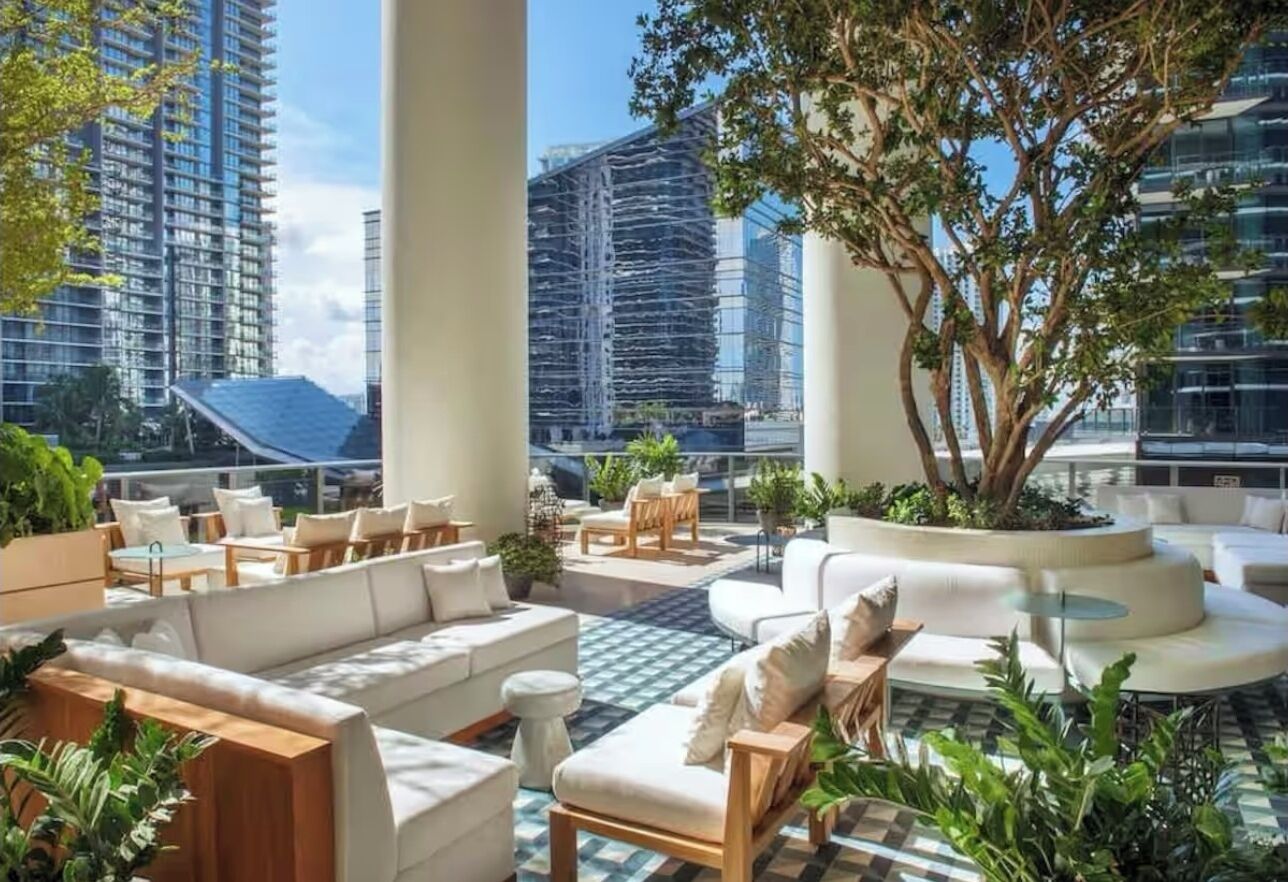 A rooftop lounge with outdoor seating, large windows, and a view of surrounding buildings.