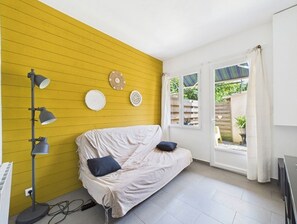 A room with a yellow wall, a covered sofa, a floor lamp, and a window with curtains. - The Musette Nest (Avignon)