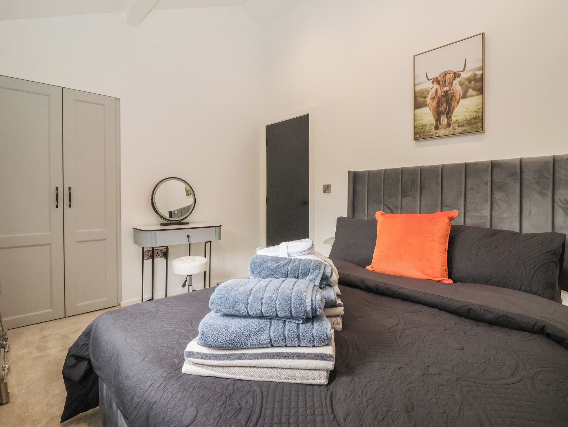 Un dormitorio con cama, una mesita de noche con espejo, un armario gris y un cuadro enmarcado en la pared.