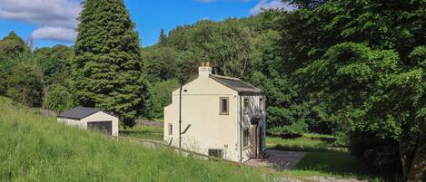 A small, single-story building with a sloped roof, surrounded by lush greenery and trees.