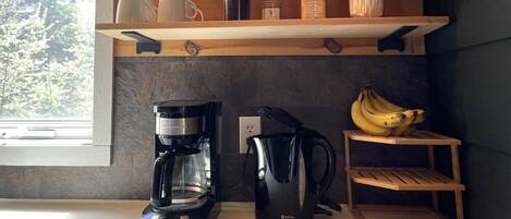 Une cuisine équipée d’une machine à café, d’une bouilloire et d’une étagère où sont rangés des bocaux et une tasse rouge.