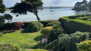Un jardín ajardinado con un gran árbol, setos cuidadosamente recortados y vistas a una masa de agua tranquila con barcos.